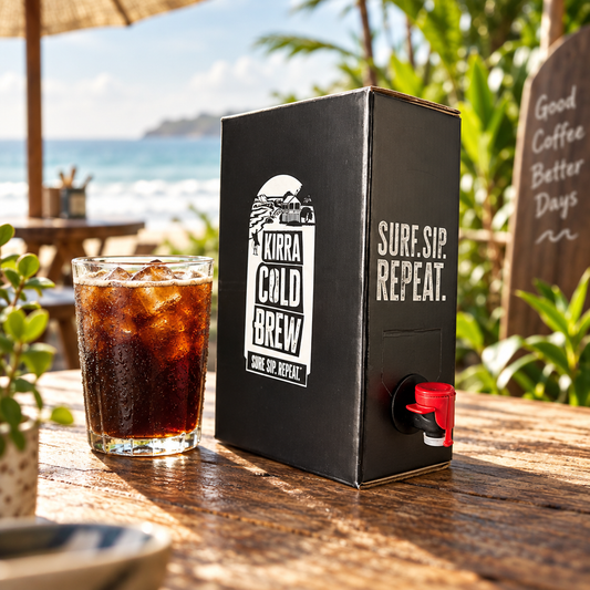 Kirra Cold Brew Coffee 1 Litre Box standing on a rustic wooden table at a beachside cafe beside a glass of iced cold brew, with the ocean and greenery behind