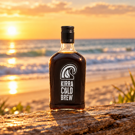 Kirra Cold Brew Coffee 350ml bottle standing on weathered timber on a Gold Coast beach at golden hour, with ocean waves and clear sunset sky behind